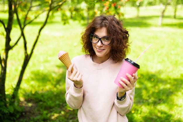 Un retrato de una mujer caucásica joven con ojos verdes, sonrisa perfecta, labios regordetes, gafas camina en la naturaleza y bebe café o cóctel y come un helado