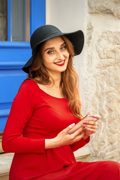 Retrato de mujer caucásica joven turista en un vestido largo rojo y sombrero negro con maleta sentado en las escaleras al aire libre.