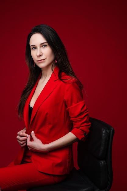 Retrato de mujer caucásica con largo cabello lacio oscuro en traje rojo de oficina, zapatos negros se sienta en una silla negra