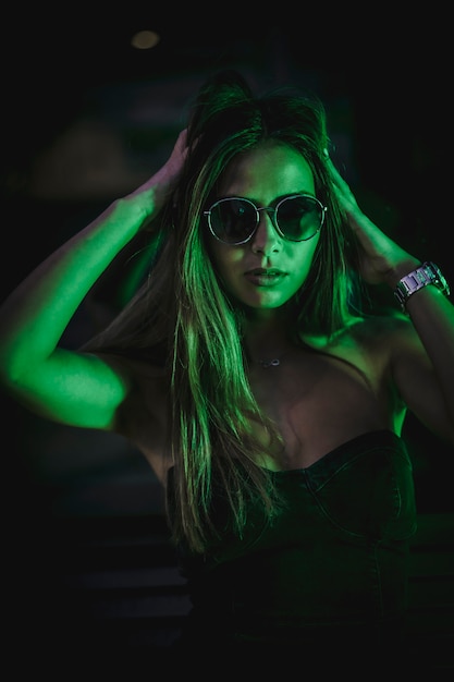 Retrato de mujer caucásica morena con gafas de sol mirando un leds verde claro. Fotografía urbana nocturna