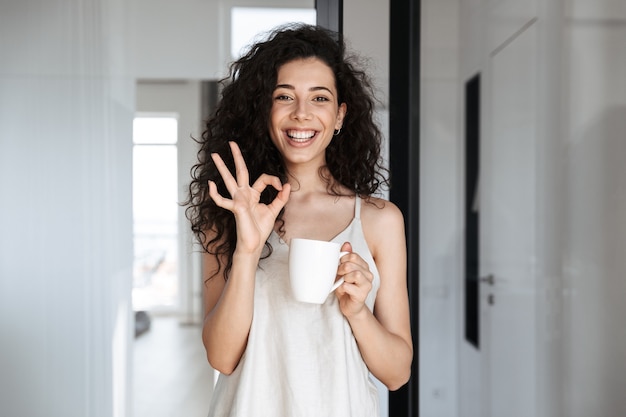 Retrato de mujer caucásica rizada con cabello largo oscuro sonriendo y mostrando el signo de ok, mientras bebe té en el apartamento del hotel o en el piso por la mañana