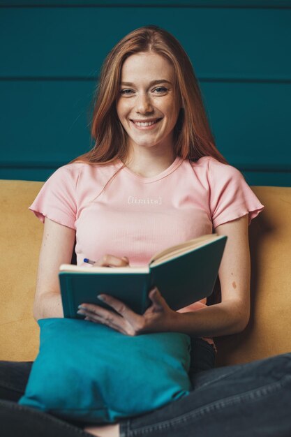 Retrato de una mujer caucásica sentada en el sofá mirando una sonrisa a la cámara pensando y escribiendo una nota...