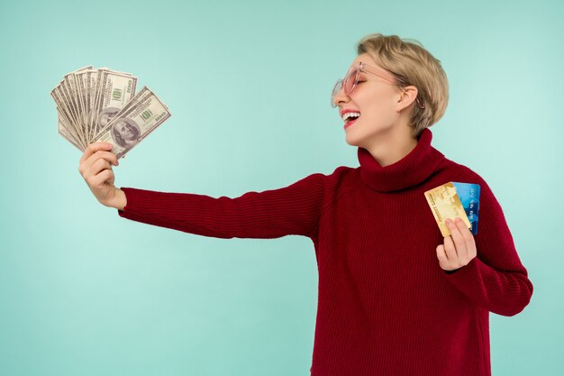 Retrato de mujer caucásica sonriente feliz con tarjeta de crédito y dinero de dólares estadounidenses mientras mira a cámara aislada sobre fondo azul.