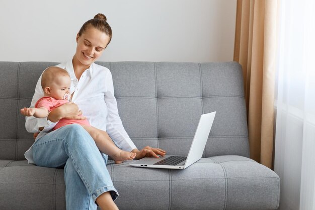 Retrato de una mujer caucásica sonriente con peinado de moño con camisa blanca y jeans sentada en la tos sosteniendo una laptop y mirando a su hija con una sonrisa