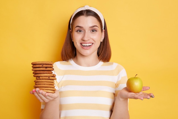 Retrato de una mujer caucásica sorprendida y emocionada que usa camiseta y banda para el cabello sosteniendo galletas y manzana prefiere fruta fresca mirando la cámara con una sonrisa dentuda posando aislada en un fondo amarillo