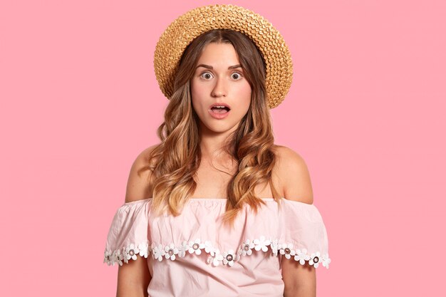 El retrato de una mujer caucásica sorprendida usa un sombrero de paja y una blusa de moda, muestra los hombros desnudos, aislados sobre la pared rosa, sorprendida de haber arruinado las vacaciones. Omg y concepto de reacción.