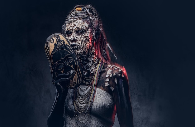 Retrato de una mujer chamán africana aterradora con una piel agrietada petrificada y dreadlocks, sostiene una máscara tradicional sobre un fondo oscuro. Concepto de maquillaje.