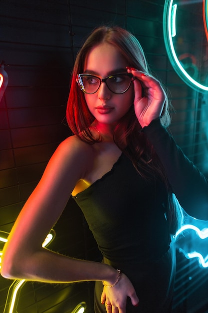 Retrato de mujer de color nocturno de una hermosa mujer de moda con ropa negra de moda con gafas elegantes en un fondo oscuro con luz de neón roja