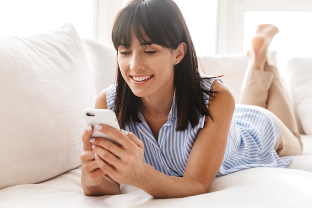 Retrato de mujer complacida con cabello oscuro sosteniendo y usando teléfono móvil mientras está acostado en el sofá en apartamento luminoso