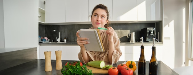 Foto retrato de una mujer comprobando notas de recetas en un cuaderno de pie en la cocina con verduras cocinando comida