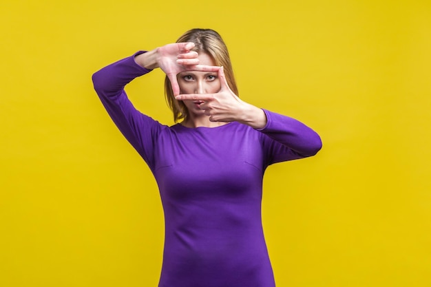 Retrato de una mujer concentrada con un elegante vestido morado ajustado mirando la cámara a través del marco de la mano, capturando una foto o enfocando la vista en el objetivo. tiro de estudio interior aislado sobre fondo amarillo