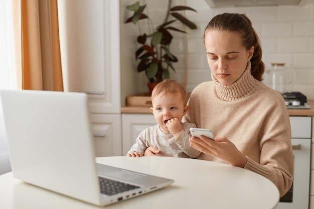 Retrato de una mujer concentrada con suéter beige sentada en la cocina y trabajando en línea en una laptop y usando el teléfono cuidando a una hija que tiene una expresión seria