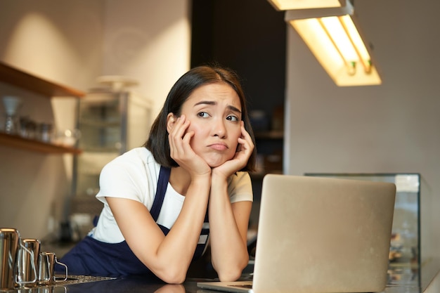 Retrato de una mujer coreana barista en un café que se ve triste y frunciendo el ceño haciendo una mueca decepcionada mientras trabaja