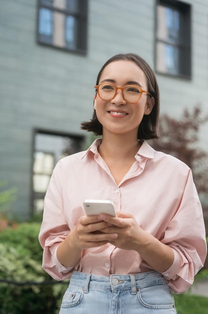 Foto retrato de una mujer coreana sonriente con anteojos sosteniendo un teléfono móvil mirando hacia otro lado en la calle