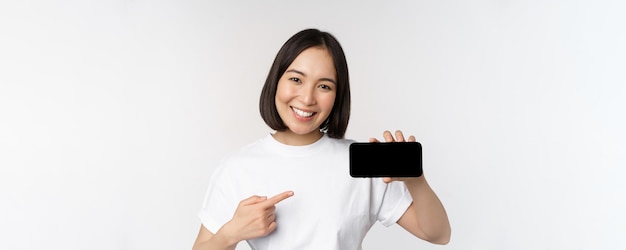 Retrato de una mujer coreana sonriente señalando con el dedo la pantalla del teléfono móvil que muestra la pantalla horizontal del teléfono inteligente recomendando el sitio web o la tienda en línea de fondo blanco