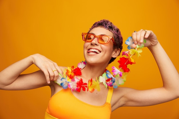 Retrato de una mujer con una corona floral hawaiana alrededor de su cuello se divierte bailando y sonriendo con ropa brillante en un fondo naranja con gafas de sol estilo hawaiano