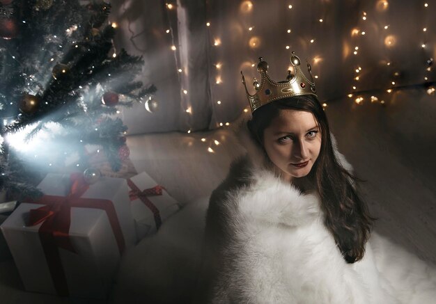 Foto retrato de una mujer con una corona mientras está sentada contra un árbol de navidad iluminado durante el invierno