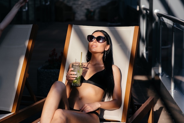 Retrato de mujer con cuerpo en forma de bronceado perfecto con gafas de sol de moda tomando un cóctel