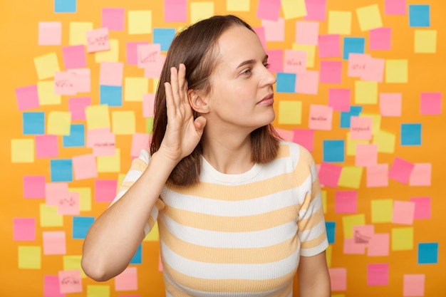 Retrato de mujer curiosa con camiseta a rayas posando sobre notas adhesivas para escribir un recordatorio en la pared amarilla de pie con la mano cerca del oído escuchando susurros