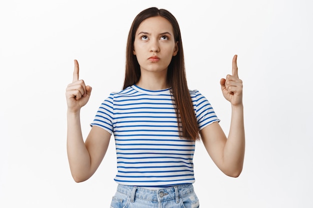 Foto retrato de mujer decepcionada señalando, mirando hacia arriba con cara molesta con el ceño fruncido molesto, quejándose, no me gusta algo malo, quejándose en un anuncio, pared blanca.