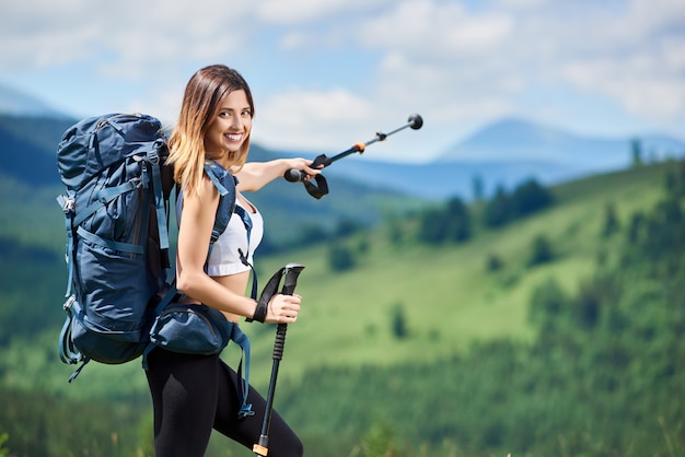 Retrato de mujer deportiva feliz turista con mochila azul apuntando lejos con sus bastones de trekking
