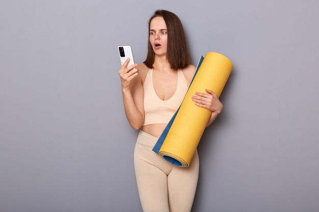 Retrato de una mujer deportiva triste y desesperada con chándal beige sosteniendo una alfombra de yoga aislada en un fondo gris usando mensajes de lectura de teléfonos móviles escuchando malas noticias