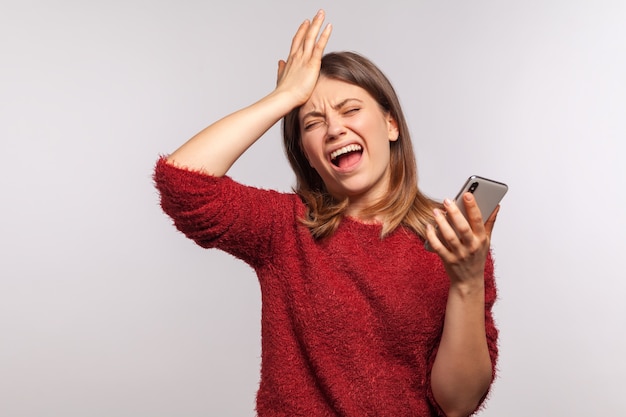 Retrato de mujer deprimida molesta de pie con gesto facepalm y sosteniendo el teléfono móvil