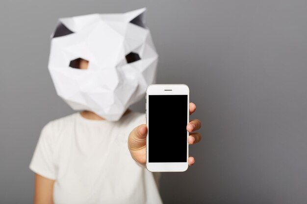 Retrato de una mujer desconocida con máscara de papel de gato y camiseta blanca que muestra la pantalla vacía del teléfono celular para el espacio de copia de publicidad para el texto promocional posando aislado sobre fondo gris