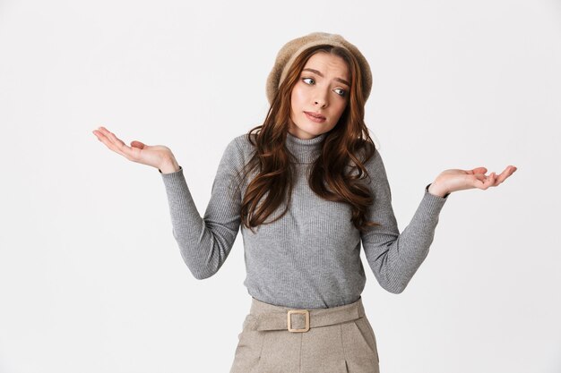 Retrato de mujer descontenta con sombrero encogiéndose de hombros y levantando las manos con impotencia aislado sobre pared blanca