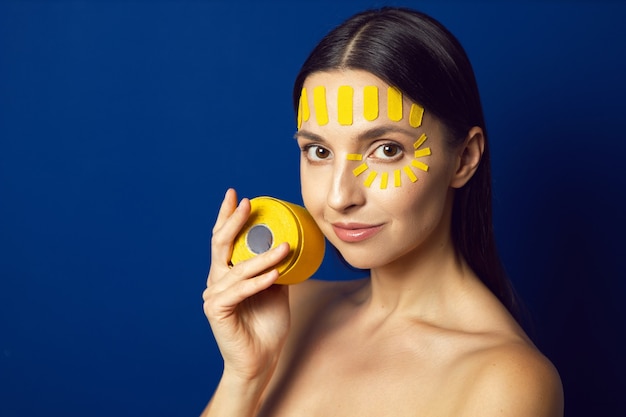 Retrato de una mujer desnuda sobre una pared azul con una cinta de kinesiotape de la cara en la frente sostiene un rollo de cinta de kinesiología en la mano