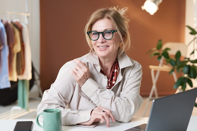 Retrato de mujer diseñadora en anteojos mirando a la cámara mientras trabaja en su lugar de trabajo en la oficina