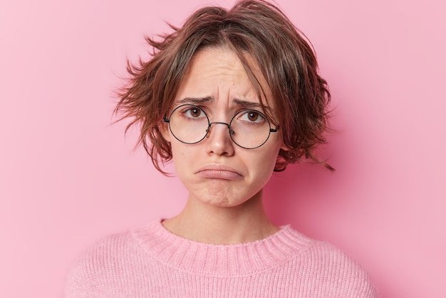 El retrato de una mujer disgustada y malhumorada se ve tristemente, los labios de los bolsos se sienten infelices y frustrados, descubre que las malas noticias usan gafas redondas, un puente suave aislado sobre un fondo rosado. Emociones negativas