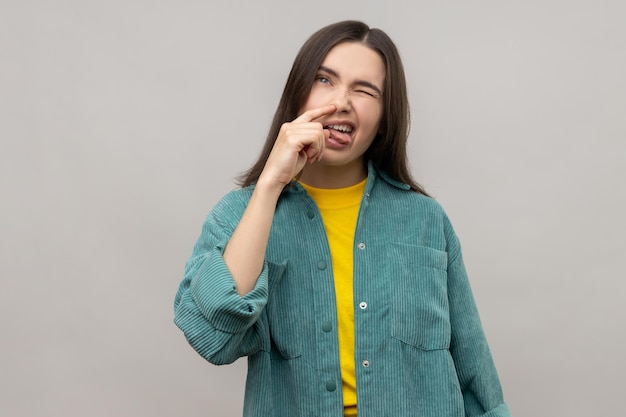 Retrato de una mujer divertida poniendo el dedo en la nariz y mostrando la lengua engañando a los malos hábitos comportamiento irrespetuoso usando una chaqueta de estilo casual Foto de estudio interior aislada en fondo gris