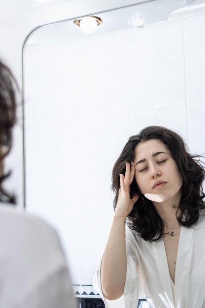 Retrato de una mujer con dolor de cabeza frente al espejo del baño en casa tocándose la cabeza