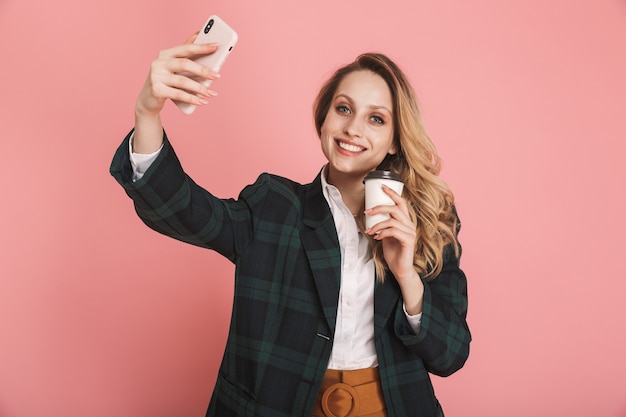 Retrato de mujer elegante de 30 años en chaqueta de moda con teléfono móvil y beber café para llevar aislado en rosa