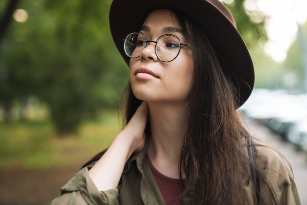 Retrato de mujer elegante hermosa con cabello largo oscuro con sombrero elegante y anteojos caminando en el parque verde