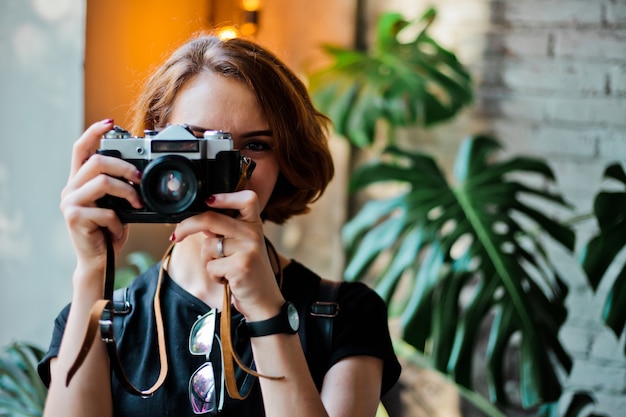 Retrato de una mujer elegante hipster con un corte de pelo corto con una cámara retro toma una foto en el interior