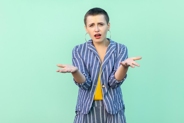 Retrato de mujer elegante joven confusa hermoso pelo corto hermoso en traje de rayas casual de pie mirando brazos levantados y pidiendo algo. tiro del estudio de interior aislado en fondo verde claro.