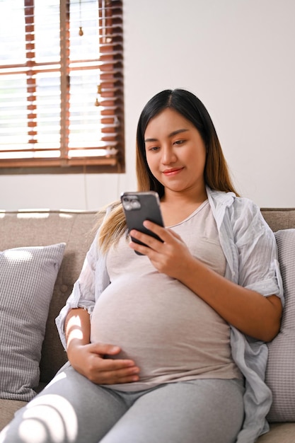 Foto retrato de una mujer embarazada asiática feliz usando su teléfono mientras se relaja en el sofá en la sala de estar