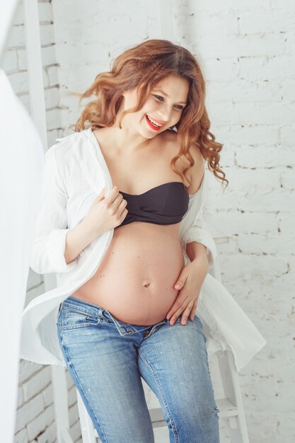 Retrato de una mujer embarazada hermosa joven feliz que se sienta en la escalera blanca. Fondo blanco