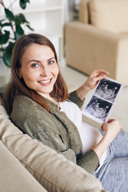 Retrato de mujer embarazada joven positiva viendo la foto de ultrasonido del bebé mientras está sentado en el sofá en casa