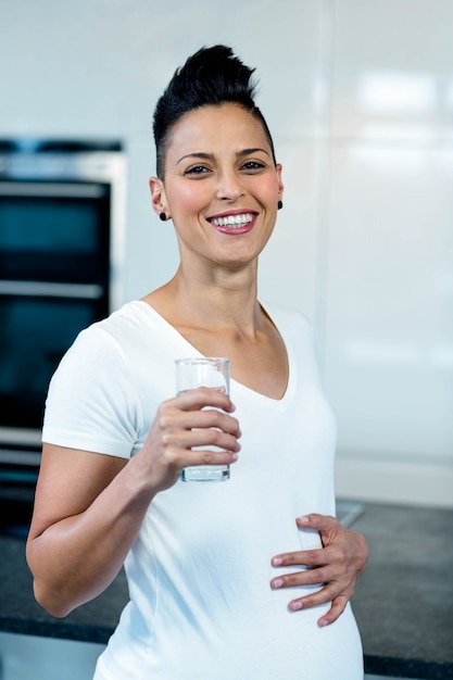 Retrato de mujer embarazada de pie con la mano sobre el estómago y el agua potable en la cocina