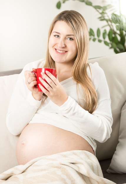 Retrato de mujer embarazada sonriente relajándose en el sofá con una taza de té