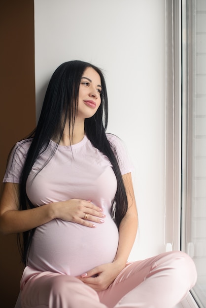 Retrato de mujer embarazada sonriente sentarse en el alféizar de la ventana