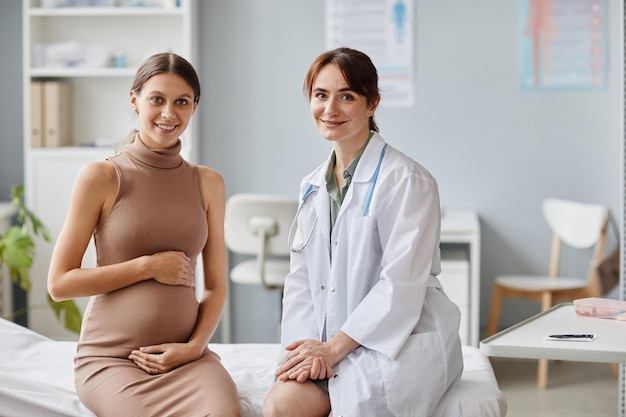 Foto retrato de una mujer embarazada y su ginecólogo sonriendo juntos a la cámara mientras se sientan en el sofá en el consultorio médico