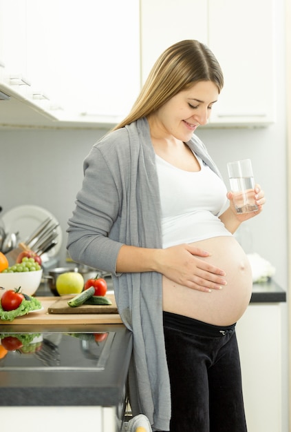 Retrato de mujer embarazada en el tercer trimestre posando con un vaso de agua en la cocina