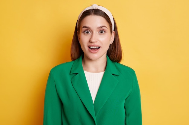 Retrato de una mujer emocionada y asombrada con una chaqueta verde y una banda para el cabello posando aislada sobre un fondo amarillo mirando la cámara con ojos grandes y boca abierta sorprendida con las noticias