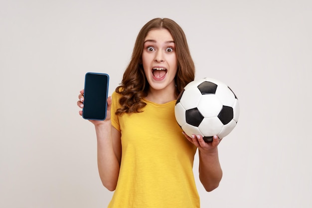 Retrato de mujer emocionada de edad joven mirando la cámara con expresión sorprendida y boca abierta, sosteniendo una pelota de fútbol y un teléfono celular con pantalla en blanco. Disparo de estudio interior aislado sobre fondo gris