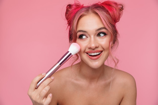 Retrato de mujer encantadora sin camisa con peinado colorido aplicando cosméticos con pincel de maquillaje