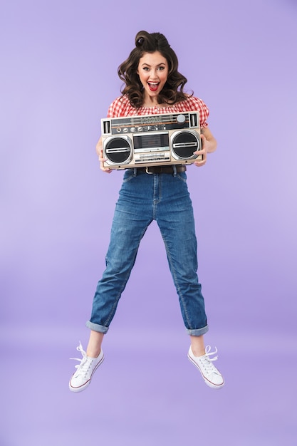 Foto retrato de mujer encantadora pin-up en estilo americano vistiendo regocijo mientras sostiene antiguo boombox vintage aislado sobre pared violeta